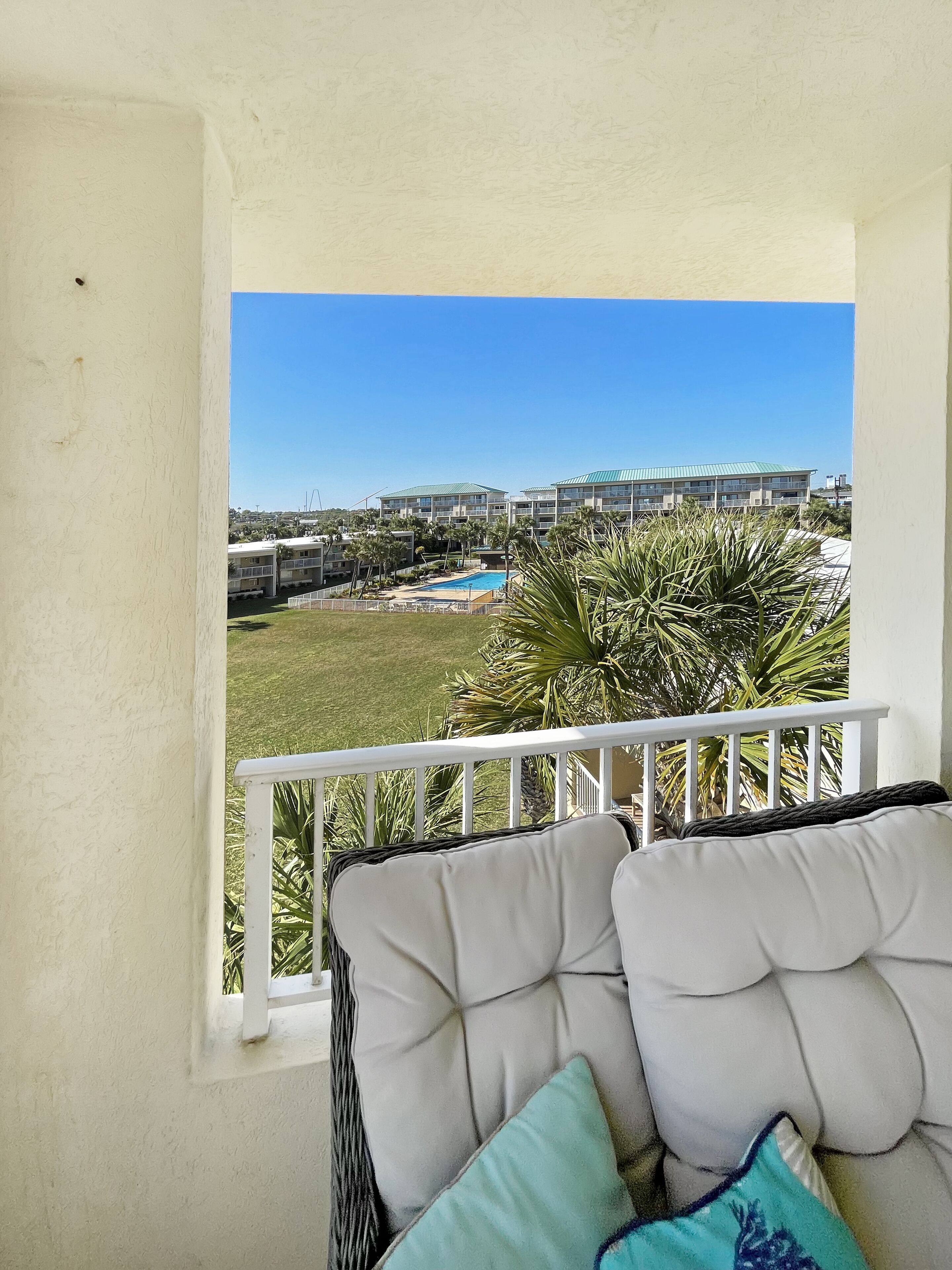 Un balcon avec des meubles en osier peints en blanc, une vue sur une piscine et, au loin, un bâtiment surmonté d’un toit végétalisé.