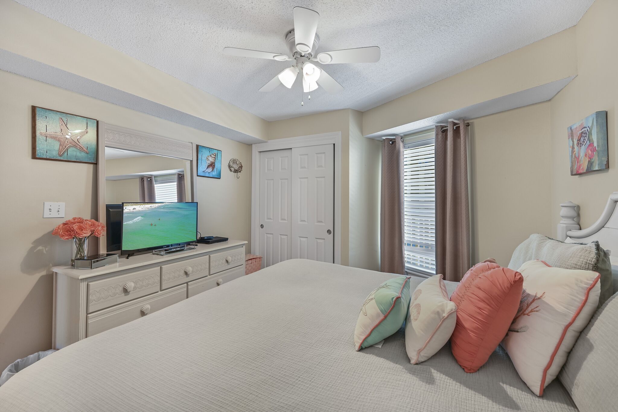 Un dormitorio con una cama, televisor, cómoda, ventilador de techo y ventana con cortinas.