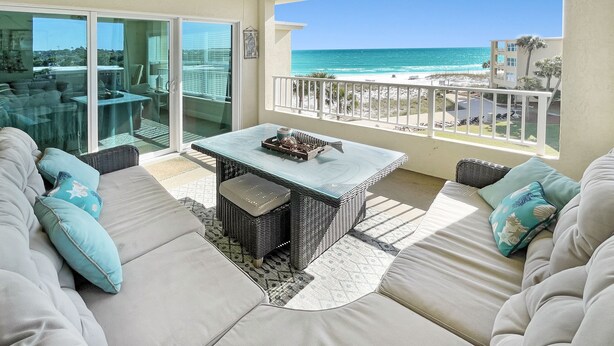A balcony with a glass door leading to a patio area, a glass table, and a sofa with cushions. - Beach-side, beautiful low-rise with uncrowded beach! (Destin)