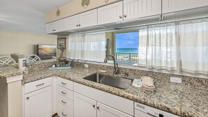A kitchen with a granite countertop, white cabinets, a stainless steel sink, and a view of the ocean through a window with sheer curtains. - Beach-side, beautiful low-rise with uncrowded beach! (Destin)