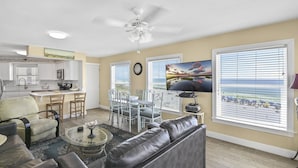 A spacious living room with a leather sofa, dining area with white chairs, a kitchen with white cabinets, and large windows with blinds.