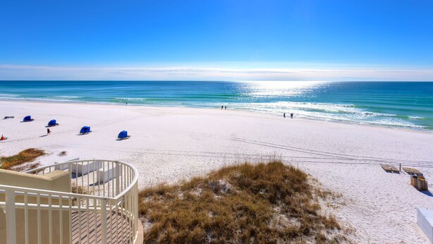 A beach with white sand, a clear blue sky, and the ocean. There are blue beach chairs and a white building with a spiral staircase.