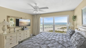 A bedroom with a large bed, a ceiling fan, a TV, a dresser, and a window with a beach view.
