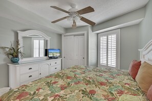 Un dormitorio con cama, ventilador de techo, cómoda, televisión y ventana con contraventanas.