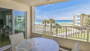 A balcony with a glass table and chairs, overlooking a beach with palm trees and a clear sky. - Beach-side, beautiful low-rise with uncrowded beach! (Destin)