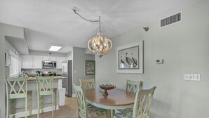 A dining area with a round table, four chairs, a chandelier, and a kitchen in the background. - Beach-side, beautiful low-rise with uncrowded beach! (Destin)