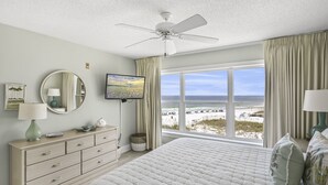 A bedroom with a large bed, a ceiling fan, a TV, a dresser, and a view of the beach.