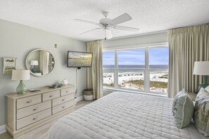 A bedroom with a large bed, a ceiling fan, a TV, a dresser, and a view of the beach.