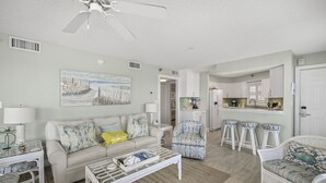 A living room with a sofa, coffee table, and chairs, a kitchen area with bar stools, and a ceiling fan.