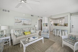 A living room with a sofa, coffee table, and chairs, a kitchen area with bar stools, and a ceiling fan.
