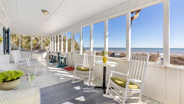 Un espace extérieur aménagé avec des sièges, protégé du soleil, doté de mobilier blanc, d’une petite table avec un pot jaune, et offrant une vue sur la plage.