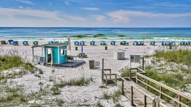 Une plage avec des parasols bleus, un poste de surveillance des sauveteurs et une promenade en bois.