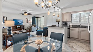 A modern living room with a glass dining table, blue chairs, a chandelier, and a ceiling fan.