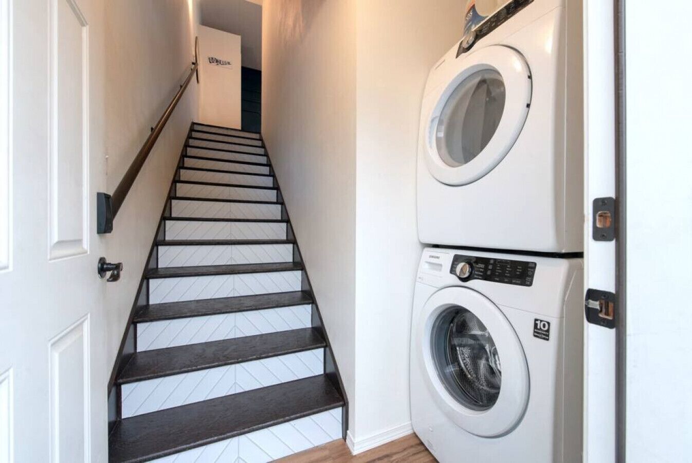 A laundry room with a washing machine and dryer, a staircase with dark wooden steps, and a white door at the top.