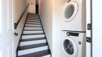 A laundry room with a washing machine and dryer, a staircase with dark wooden steps, and a white door at the top.