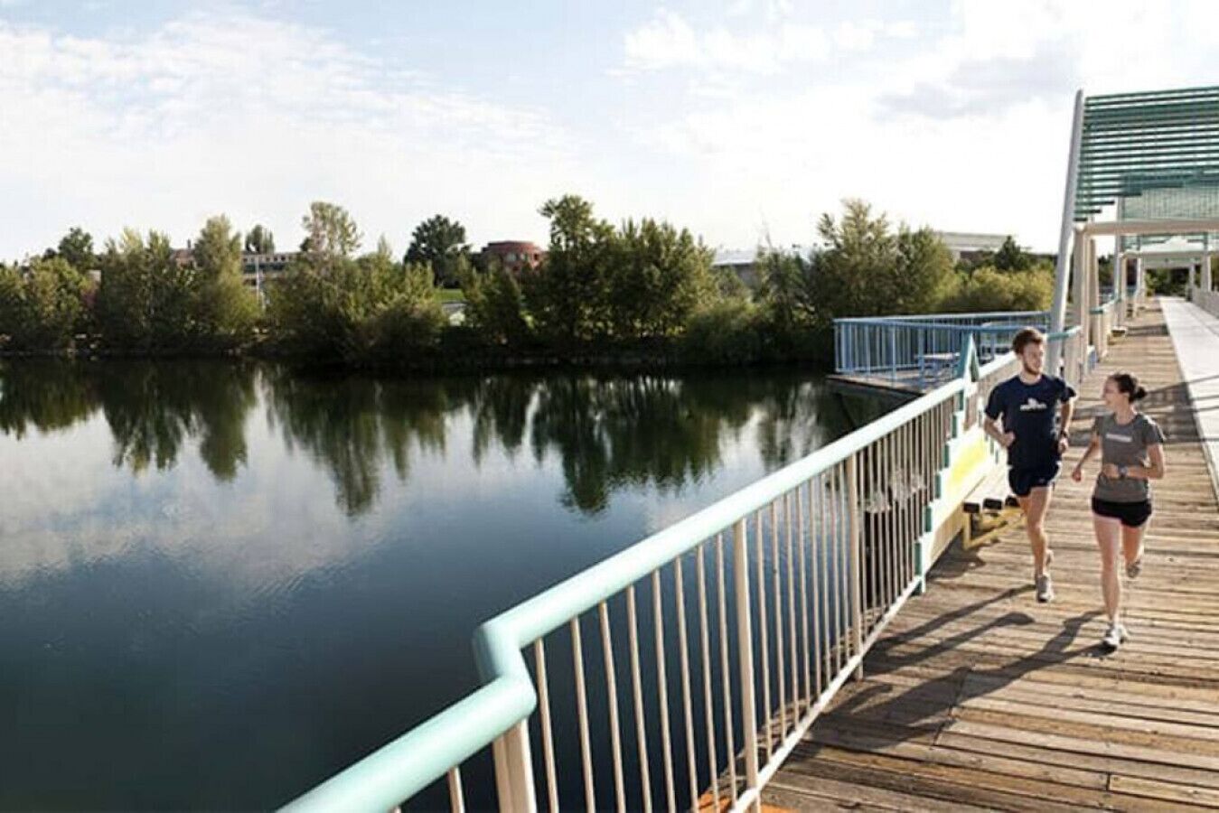 A wooden boardwalk with a metal railing overlooking a calm body of water, surrounded by trees and buildings in the background.