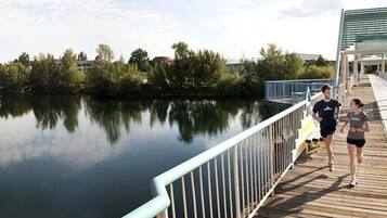 A wooden boardwalk with a metal railing overlooking a calm body of water, surrounded by trees and buildings in the background.