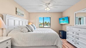 A bedroom with a bed, a ceiling fan, a TV, a dresser, and a window with a view of palm trees and a beach.