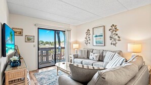 Un salon avec un canapé, une table basse et un téléviseur. Il y a un balcon avec vue sur des palmiers et une plage.
