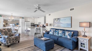 Uma sala de estar moderna com um sofá azul, poltrona estampada e um banquinho. Há uma área de jantar com um lustre e uma cozinha ao fundo.