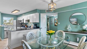 A modern kitchen with white cabinets, a central island, and a dining area with a glass table and white chairs. - Newly updated!! 2 Bed 2 Bath overlooking the pool and courtyard. (Destin)