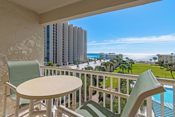 A balcony with a table and chairs, overlooking a resort area with a pool and ocean view.