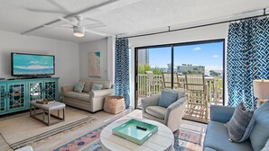 A modern living room with a sofa, armchair, and a coffee table. There is a television on a cabinet, and a balcony with a view of buildings. - Beach-side, beautiful low-rise with uncrowded beach! (Destin)