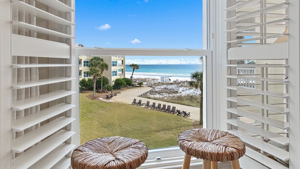 Ein Balkon mit weißen Geländern und Korbstühlen, der einen Blick auf einen Strand mit Liegestühlen und ein Gebäude im Hintergrund freigibt.