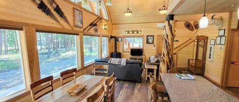 Interior de una cabaña de madera con comedor, asientos, televisor y ventanas amplias.