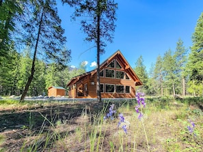 Puinen mökki, jossa on suuri ikkunallinen etuosa, ja sen ympärillä on tiheää metsää sekä pieni varasto.