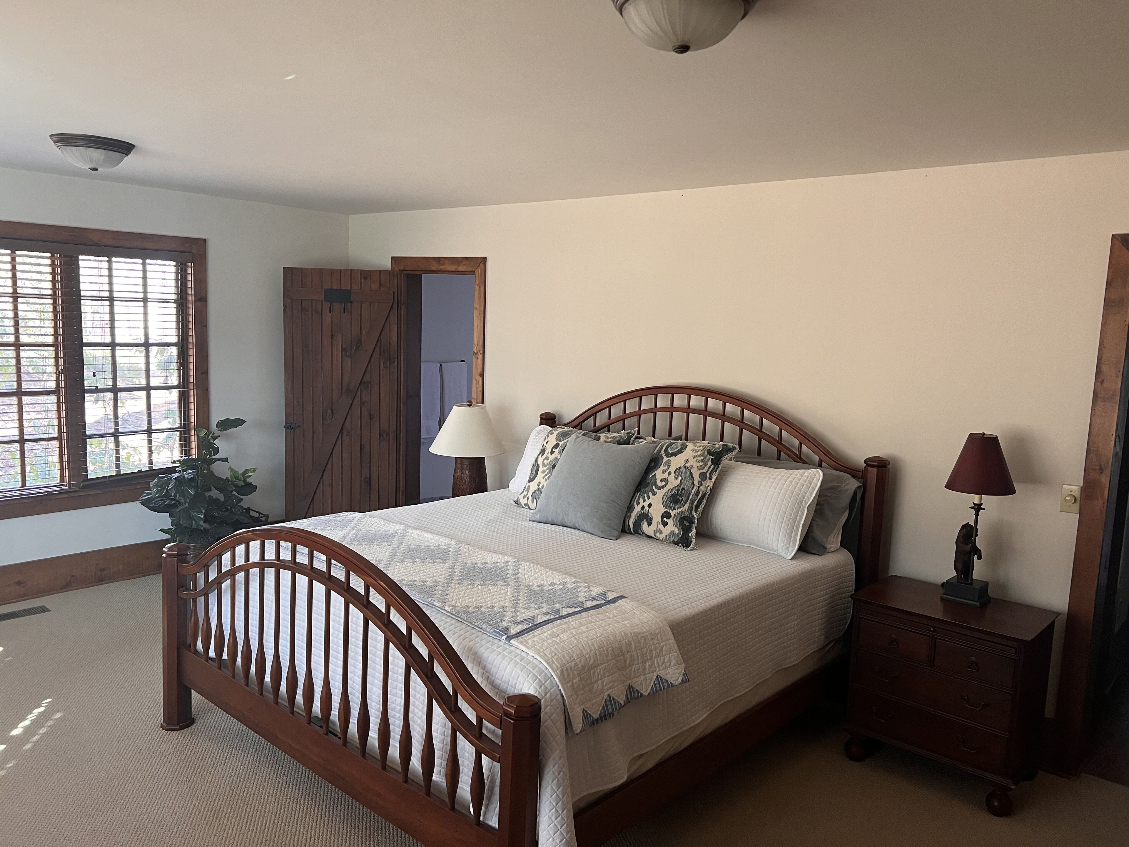 A bedroom with a wooden bed, a nightstand, a lamp, a window with blinds, and a door.