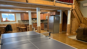 A ping pong table in a room with wooden flooring, a kitchen area with wooden cabinets, a refrigerator, and a staircase.