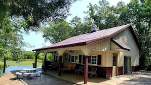 A covered outdoor seating area with picnic tables and chairs, a covered porch, and a view of a lake.