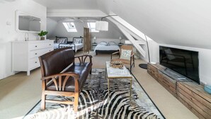 A spacious loft bedroom with a large bed, a seating area with a leather sofa, a TV, and a view of the city through skylights.