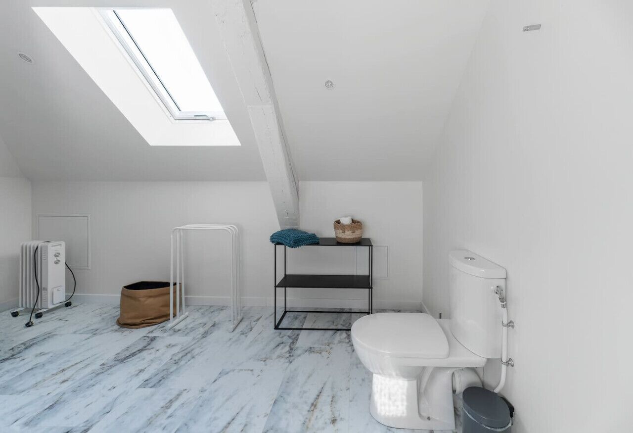 Een kleine, witte badkamer in zolderstijl met dakraam, een wit toilet, een radiator en een metalen plankje met handdoek en mandje.