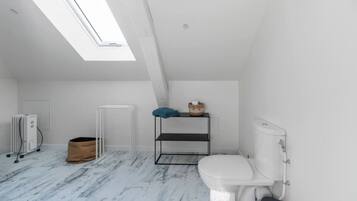 Een kleine, witte badkamer in zolderstijl met dakraam, een wit toilet, een radiator en een metalen plankje met handdoek en mandje.