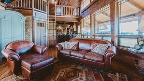 A wooden interior with a brown leather sofa and armchair, a patterned rug, and a view through a large window. - The Retreat at Rusty Elk Lodge, Star Valley Wyoming (Thayne)