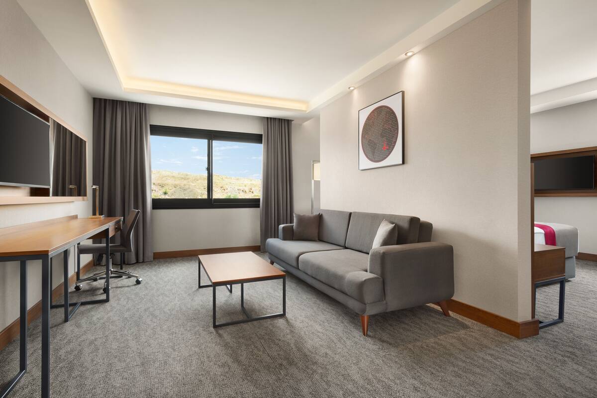 Habitación de hotel moderna con sofá, una mesita, escritorio y un amplio ventanal con vistas al desierto.