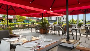 Un área de comedor al aire libre con mesas dispuestas para dos, sombrillas rojas y vistas de los edificios circundantes.