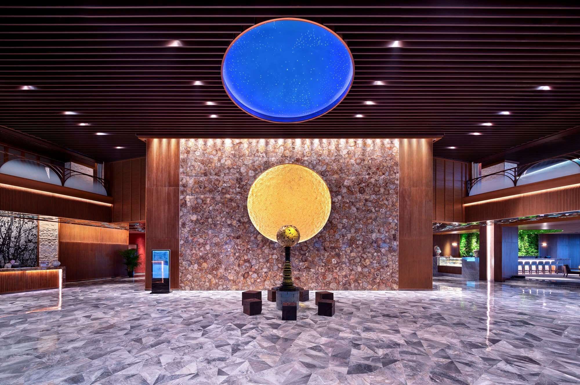 A spacious lobby with a large, circular, illuminated ceiling feature, a textured stone wall, and a central sculpture.