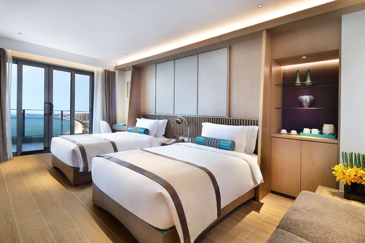 A hotel room with two beds, a large window with a view, a small desk, and a shelf with decorative items.