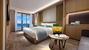 A hotel room with a large bed, a small table with a vase of flowers, a sofa, and a view of the sea through the glass door. - Wyndham Grand Plaza Royale Shuangyue Bay (Huizhou)