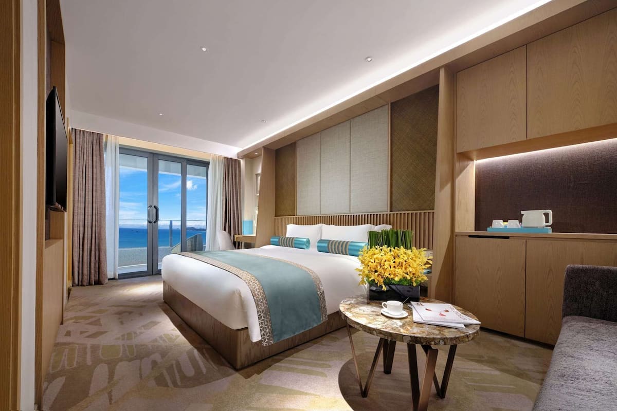A hotel room with a large bed, a small table with a vase of flowers, a sofa, and a view of the sea through the glass door.