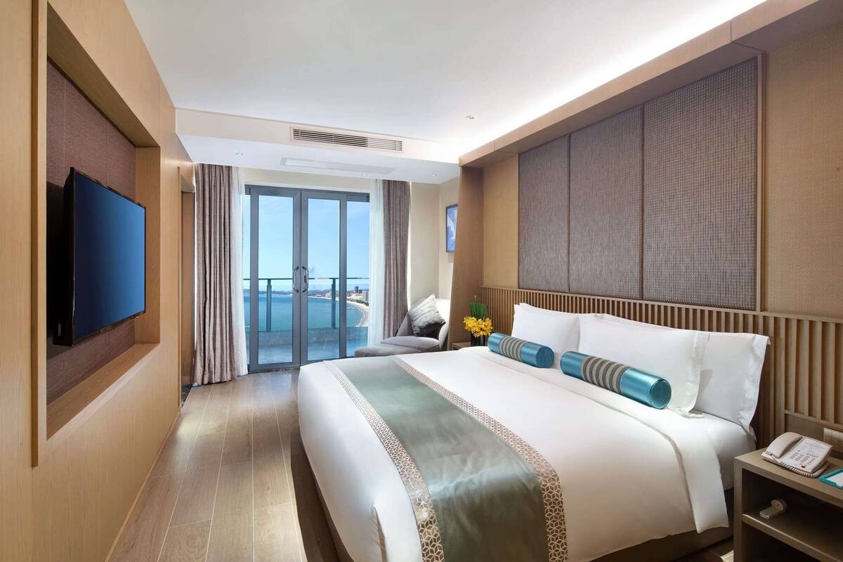 A hotel room with a large bed, a flat-screen TV, a balcony with a view of the sea, and a large window.
