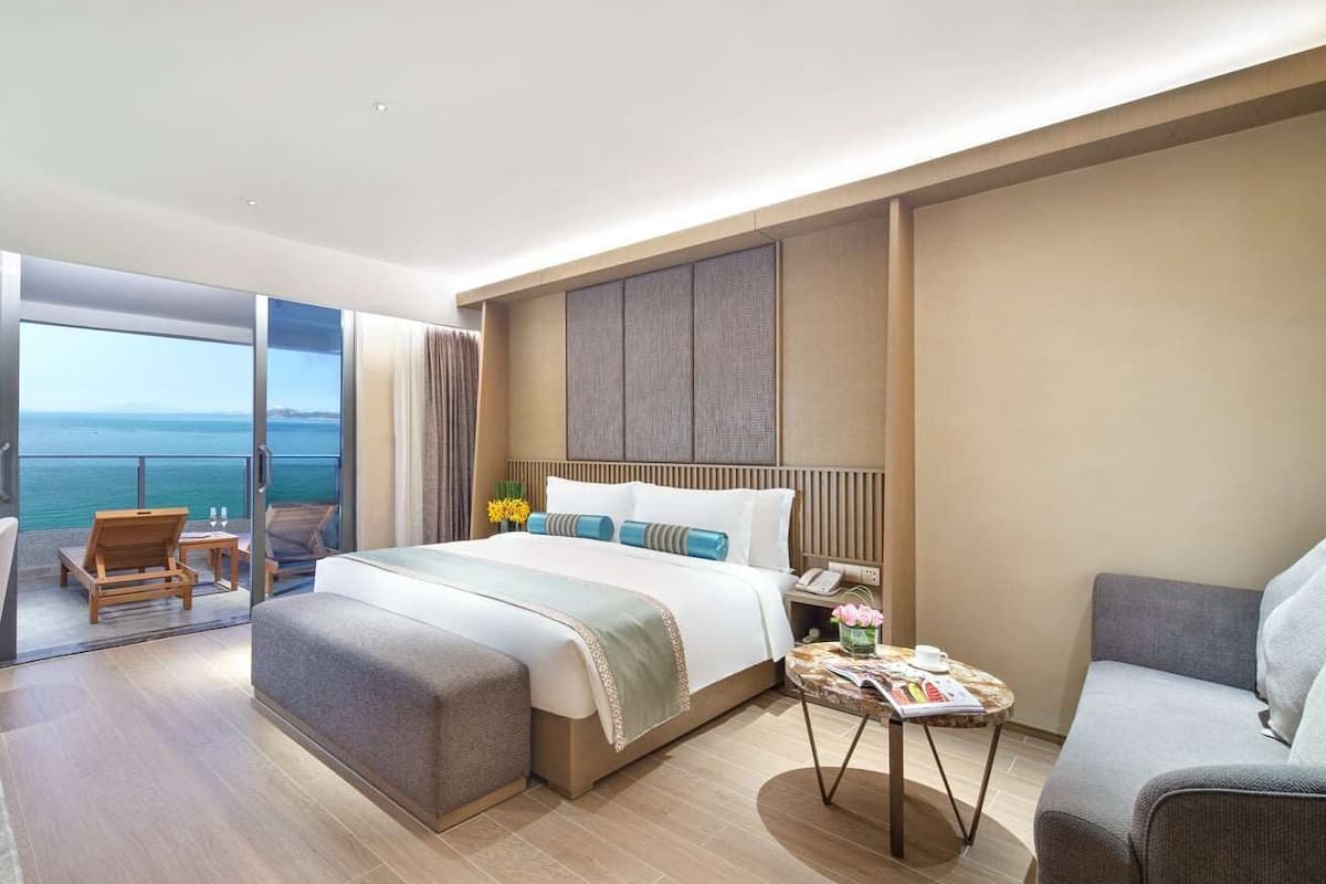 A modern hotel room with a large bed, a sofa, a small table, and a balcony with a view of the sea.