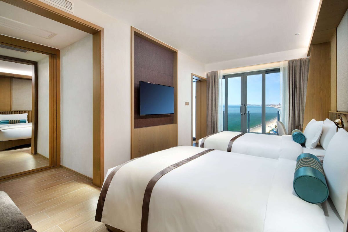 A hotel room with two beds, a TV, and a large window with a view of the sea.