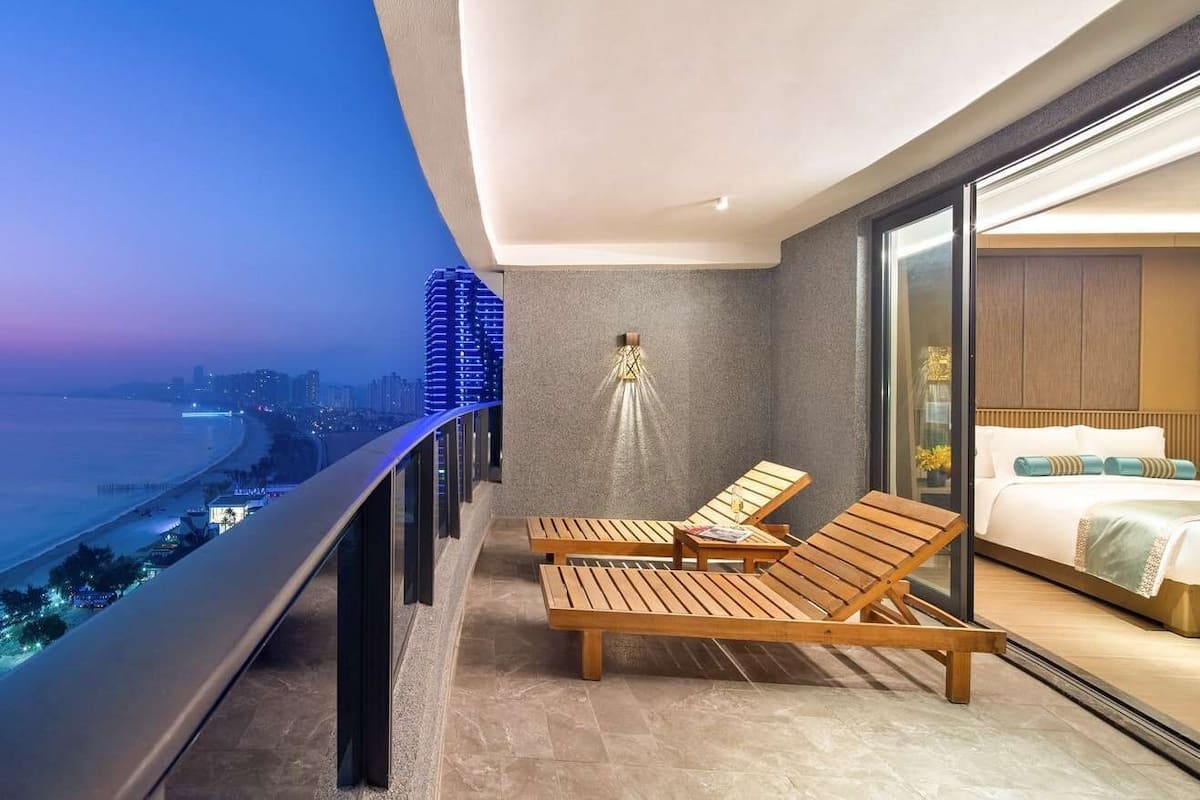 A balcony with two wooden lounge chairs, a small table, and a view of a cityscape at dusk.