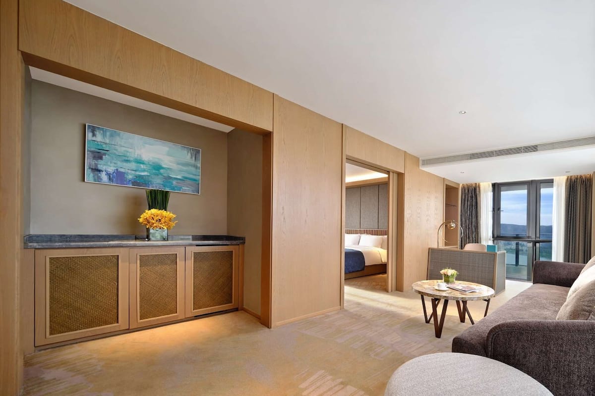 A modern hotel room with a wooden cabinet, a painting, a vase with flowers, a bed, a seating area with a table, and a view of the outdoors.