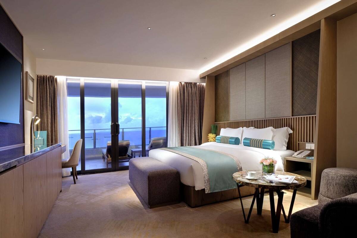 A modern hotel room with a large bed, a small round table, a seating area with a chair, and a view of the sea through large windows.