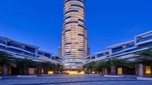 A modern hotel with a cylindrical tower, multiple balconies, and a spacious entrance area with palm trees. - Wyndham Grand Plaza Royale Shuangyue Bay (Huizhou)